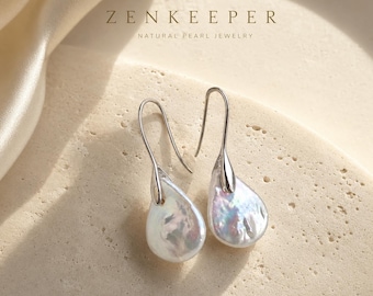 Puede incluir: Un par de pendientes colgantes con perlas barrocas iridiscentes en forma de lágrima suspendidas de ganchos de metal plateado. El texto ZENKEEPER NATURAL PEARL JEWELRY es visible sobre los pendientes, expuestos en una superficie texturizada.