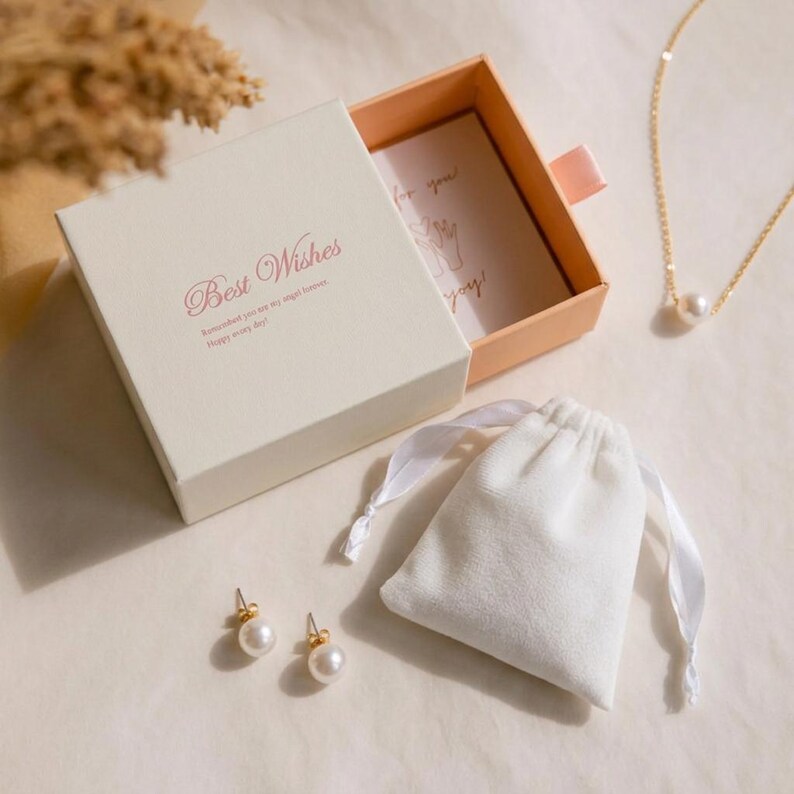 Puede incluir: Una caja de regalo cuadrada de color crema con el texto Best Wishes impreso en rosa, un caj&oacute;n abierto de color melocot&oacute;n con una tarjeta, una bolsa de terciopelo blanco con cord&oacute;n, dos pendientes de perlas y una cadena dorada con perla.
