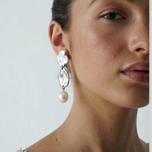Peut inclure: Une boucle d'oreille pendante argentée composée de deux plaques métalliques aux formes organiques irrégulières superposées verticalement, avec une perle ronde blanc cassé suspendue en bas. La surface est polie et réfléchissante.