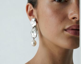 Puede incluir: Un pendiente colgante de tono plateado con dos placas de metal de formas orgánicas irregulares apiladas verticalmente, con una perla redonda de color blanco roto suspendida en la parte inferior. La superficie es pulida y reflectante.