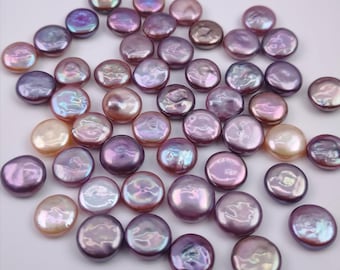 Puede incluir: Una colección de perlas sueltas, redondas y planas en forma de moneda, en varios tonos de morado, lavanda y rosa suave. Cada perla presenta una superficie lisa e iridiscente con un acabado brillante, mostrando sutiles variaciones de color.