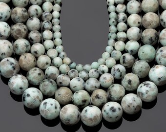 Puede incluir: Primer plano de una colección de cuentas redondas y moteadas de varios tamaños. Las cuentas son principalmente de color verde claro con manchas grises oscuras y negras, creando una apariencia natural y terrosa. Las cuentas están dispuestas en un patrón curvo.