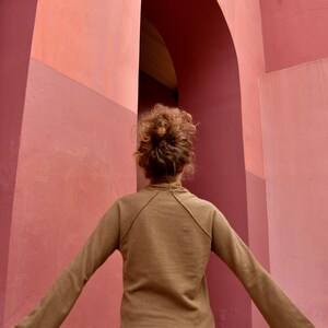 May include: A person with brown hair wearing a brown long-sleeved shirt stands in front of a pink wall with an archway.