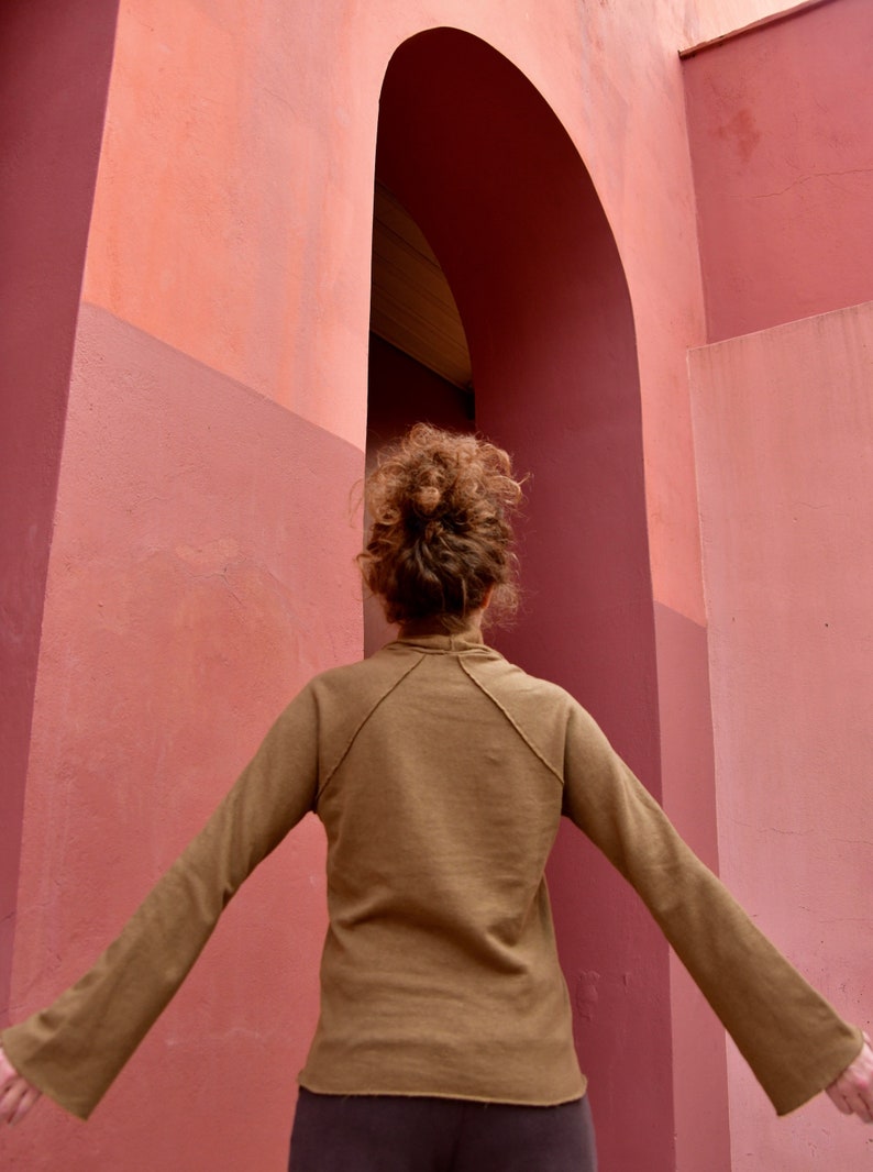 May include: A person with brown hair wearing a brown long-sleeved shirt stands in front of a pink wall with an archway.