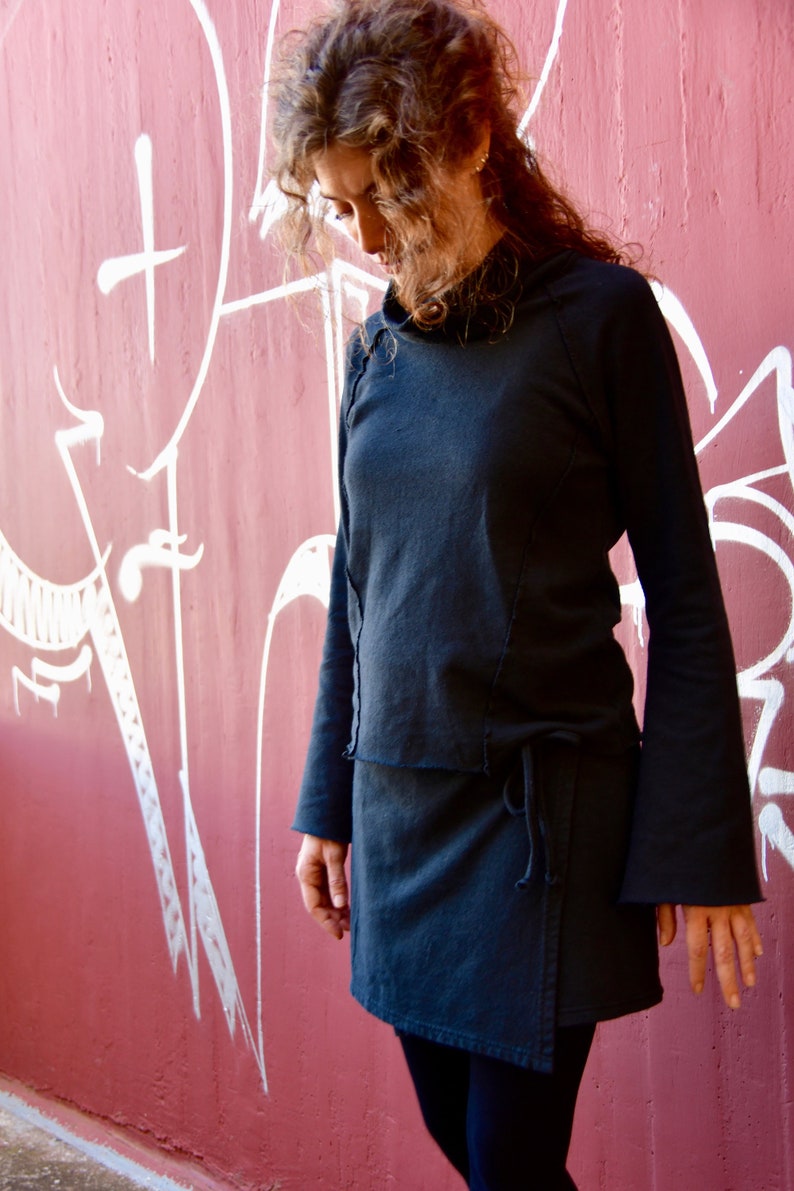 May include: A black hooded top with long sleeves and a wrap skirt. The top has a raw edge detail and the skirt has a tie closure. The person is standing in front of a red wall with white graffiti.