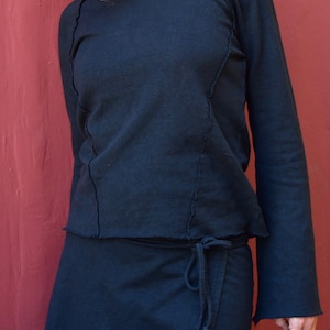 May include: A black mock neck shirt with long sleeves and a wrap skirt. The shirt has a raw edge detail and the skirt has a tie closure.