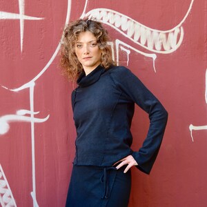 May include: A woman wearing a black mock neck shirt and a black wrap skirt. She is standing in front of a red wall with white graffiti.