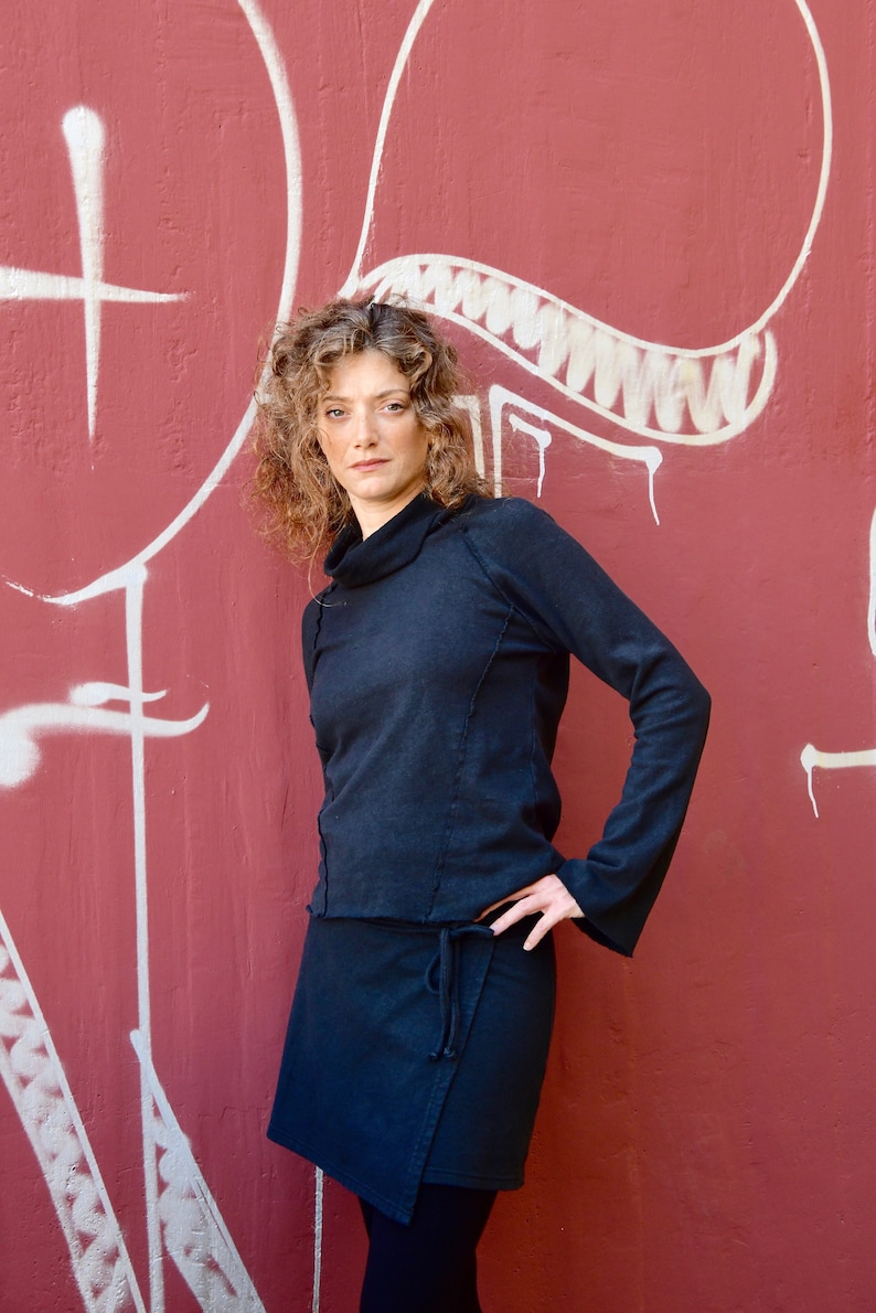 May include: A woman wearing a black mock neck shirt and a black wrap skirt. She is standing in front of a red wall with white graffiti.