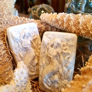 May include: Two white soap bars with a mermaid design. The soap bars are surrounded by seashells and starfish.