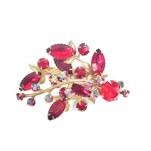 May include: A gold-toned brooch with a red rhinestone design. The brooch features a cluster of red rhinestones arranged in a leaf-like pattern.