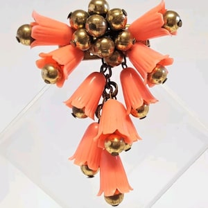 Puede incluir: Un broche vintage con un racimo de campanas de plástico de color coral con cuentas de metal dorado y una cadena. El broche es una pieza llamativa con un diseño único.
