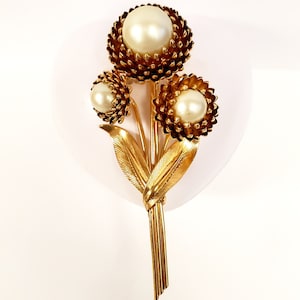 Puede incluir: Un broche dorado con tres flores de perlas falsas con un tallo de hojas doradas y un borla dorada.