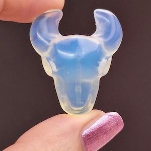 Könnte beinhalten: Ein kleiner, durchscheinender blauer und weißer Opal-Schädel-Anhänger mit Hörnern. Der Anhänger ist aus Glas und hat eine glatte, polierte Oberfläche.