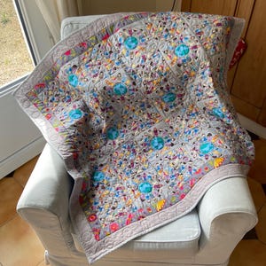 Peut inclure: Une courtepointe faite à la main avec un fond gris et un motif coloré de personnes et de globes. La courtepointe est drapée sur un fauteuil bleu clair. La bordure est grise unie avec un motif matelassé.