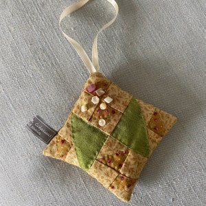Peut inclure: Une petite décoration carrée matelassée avec un motif patchwork vert et marron. Elle est dotée d'un ruban de satin blanc pour l'accrocher et est décorée de petits boutons blancs et de perles roses. La décoration porte une étiquette qui indique "Créations  Main".