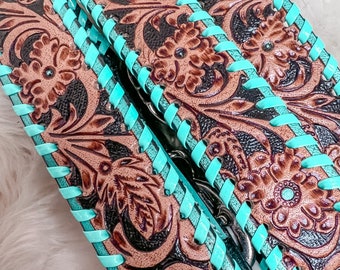 May include: Two brown leather belts with turquoise stitching and an intricate floral pattern. The belts are folded over each other, showing the stitching and pattern on both sides.