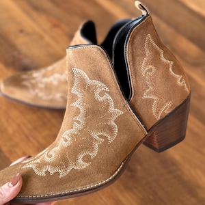 May include: Brown suede ankle boots with a cut-out detail and white embroidered floral designs. The boots have a block heel.