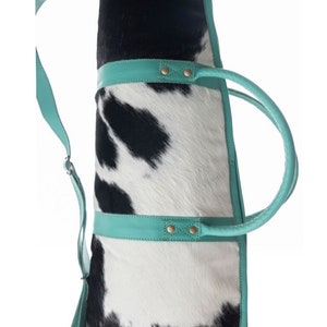 Puede incluir: Un bolso tote de piel de vaca blanco y negro con ribetes y asas de cuero turquesa. El bolso tiene una correa larga para el hombro.