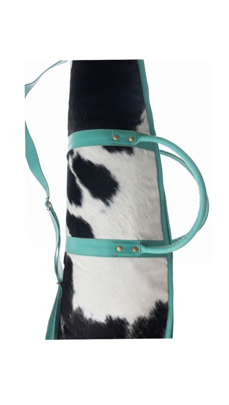 Puede incluir: Un bolso tote de piel de vaca blanco y negro con ribetes y asas de cuero turquesa. El bolso tiene una correa larga para el hombro.