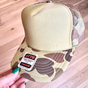 Puede incluir: Una gorra de camionero de camuflaje marrón y beige con un frente de espuma y un cierre a presión. La gorra tiene una etiqueta que dice "OTTO World Famous Foam Front OSFM Trucker Durable Snap Closure".