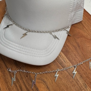 Könnte beinhalten: Zwei silberne Ketten mit Blitzscharmen. Die Ketten liegen auf einer weißen Trucker-Kappe und einer Holzoberfläche.