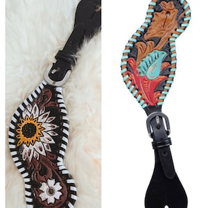 Può includere: Due cinturini per stivali in pelle nera con ricami floreali decorativi. Un cinturino presenta un motivo a girasole con accenti bianchi e gialli, mentre l'altro presenta un motivo floreale con accenti turchese, rosso e marrone.