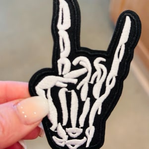 May include: Black and white embroidered patch of a skeleton hand making the rock and roll hand gesture.