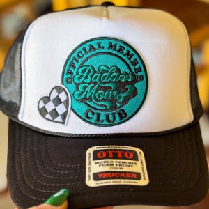 Op de afbeelding: Witte en zwarte trucker cap met een turquoise blauwe patch waarop staat "Official Member Badass Moms Club". De patch heeft een zwart-wit geruit hart aan de linkerkant.