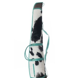 Puede incluir: Una funda para rifle de piel de vaca en blanco y negro con detalles en turquesa y una base de cuero marr&oacute;n. La funda tiene una correa para el hombro y dos asas de transporte.