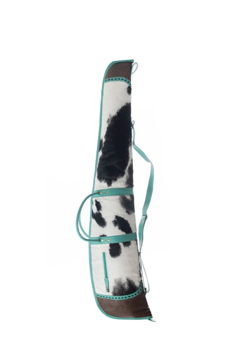 Puede incluir: Una funda para rifle de piel de vaca en blanco y negro con detalles en turquesa y una base de cuero marr&oacute;n. La funda tiene una correa para el hombro y dos asas de transporte.
