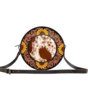 May include: A round brown leather crossbody bag with a white and brown cowhide patch in the center. The bag is decorated with a sunflower design and has a long adjustable strap.