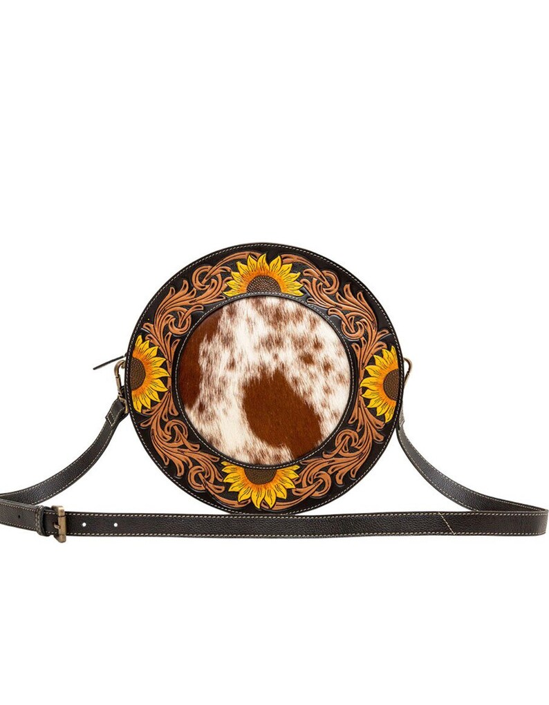May include: A round brown leather crossbody bag with a white and brown cowhide patch in the center. The bag is decorated with a sunflower design and has a long adjustable strap.