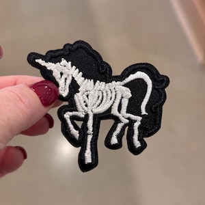 Könnte beinhalten: Schwarzer, gestickter Aufnäher mit einem weißen Einhorn-Skelett-Design. Der Aufnäher hat einen schwarzen Rand. Das Design ist detailliert und für Kleidung oder Accessoires geeignet. Der Aufnäher wird von einer Hand gehalten.