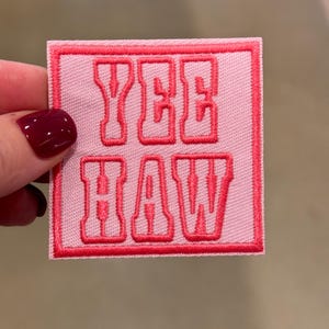 Puede incluir: Parche de tela rosa con un borde bordado rojo y las palabras "Yee Haw" en rojo.