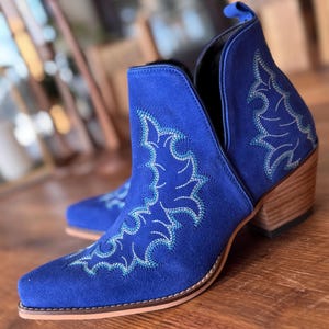May include: Blue suede ankle boots with a pointed toe and a low block heel. The boots feature a decorative embroidered design in a lighter blue color.
