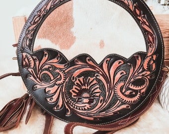May include: A round, brown leather purse with a white and brown cowhide patch in the center. The purse has intricate floral designs carved into the leather and a long brown leather strap.