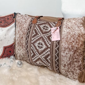 Puede incluir: Un cojín rectangular con un panel central de patrones geométricos tejidos en marrón y blanco, flanqueado por tela con estampado animal en marrón y blanco. El cojín incluye asas de aspecto cuero marrón y flecos en los bordes.
