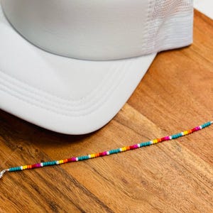 May include: A white trucker hat with a mesh back sits on a wooden surface next to a beaded bracelet. The bracelet features a colorful pattern of red, pink, yellow, orange, and teal beads.