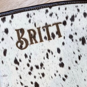 Puede incluir: Un bolso de cuero de vaca blanco y negro con el nombre "BRITT" bordado en hilo marr&oacute;n.