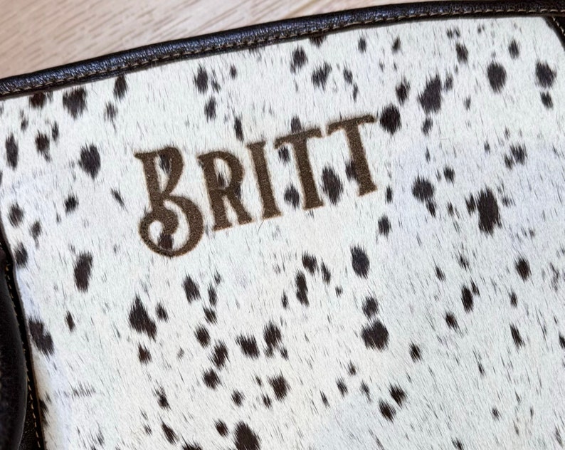 Puede incluir: Un bolso de cuero de vaca blanco y negro con el nombre "BRITT" bordado en hilo marr&oacute;n.