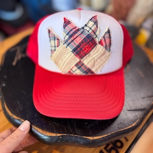 May include: A red and white trucker hat featuring a central patch made of quilted fabric squares in red, navy blue, and beige plaid patterns. The hat has a solid red brim with horizontal stitching and a white front panel with the fabric applique.