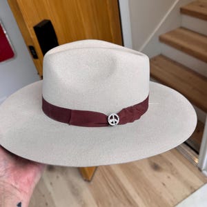 Puede incluir: Un sombrero fedora de fieltro gris claro con ala ancha y una cinta de color burdeos. El sombrero tiene un pin con un símbolo de paz en el centro de la cinta.
