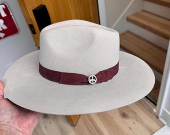 May include: A light grey felt fedora hat with a wide brim and a burgundy ribbon band. The hat has a peace sign pin in the centre of the ribbon.