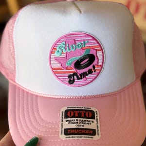 Puede incluir: Gorra de camionero rosa con un panel frontal blanco y un parche bordado rosa y blanco que dice "River Time!" en una fuente cursiva. El parche presenta un contorno estilizado del estado de Texas con un círculo turquesa en su interior. La gorra tiene una malla negra en la parte posterior y un cierre de presión negro.