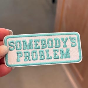 Puede incluir: Parche blanco bordado con un borde azul turquesa y el texto "Somebody's Problem" en azul turquesa.