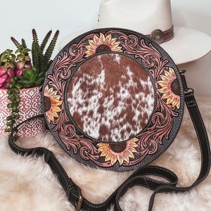 May include: A round, brown leather purse with a cowhide patch and sunflower designs. The purse has a black adjustable strap.