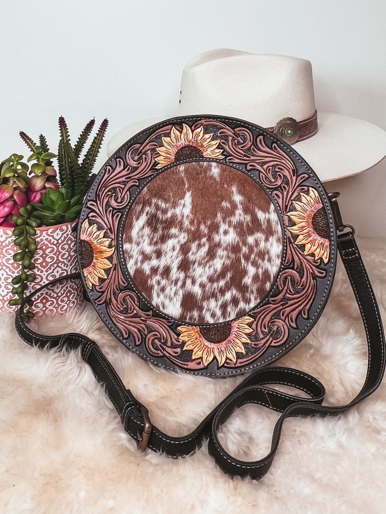 May include: A round, brown leather purse with a cowhide patch and sunflower designs. The purse has a black adjustable strap.