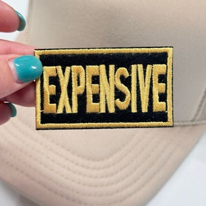 Op de afbeelding: Een geborduurde zwarte en gouden patch met het woord "EXPENSIVE" in gouden letters.