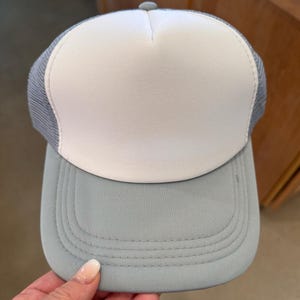 May include: A white and light grey trucker hat with a mesh back. The hat has a flat brim and a curved crown.