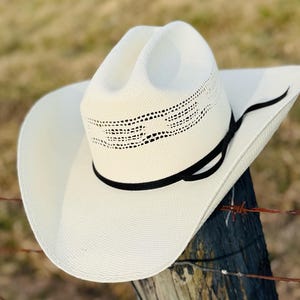 Pode incluir: Um chapéu de cowboy branco com uma faixa preta e um design tecido em torno da coroa. O chapéu está sentado em um poste de madeira.